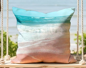 May include: A square cushion depicting a beach scene with turquoise water, white waves, and a pink sandy shore. The cushion is on a wooden swing with rope supports, set against a blurred beach background.