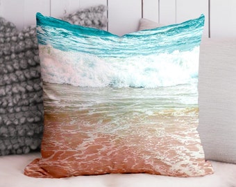May include: A decorative cushion featuring a beach scene with turquoise blue ocean waves breaking on a sandy shore. The cushion has a soft, peach-coloured background.