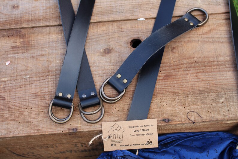 Puede incluir: Dos correas de cuero negro con anillas en D de lat&oacute;n y una etiqueta de papel marr&oacute;n que dice "Sangle de sac fourre-tout, Long 120 cm, Cuir/Tannage v&eacute;g&eacute;tal, Fabriqu&eacute; en Afrique par Jo."