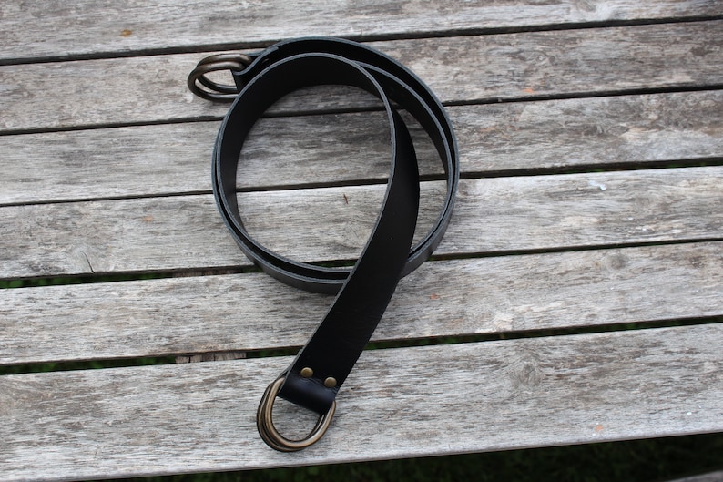 Puede incluir: Un cintur&oacute;n de cuero negro con una hebilla de doble anillo de lat&oacute;n.