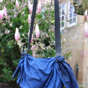 Puede incluir: Una bolsa de tela azul con un patr&oacute;n de hojas que cuelga de correas de cuero negro. La bolsa est&aacute; atada en la parte superior con un nudo y tiene un aspecto informal.