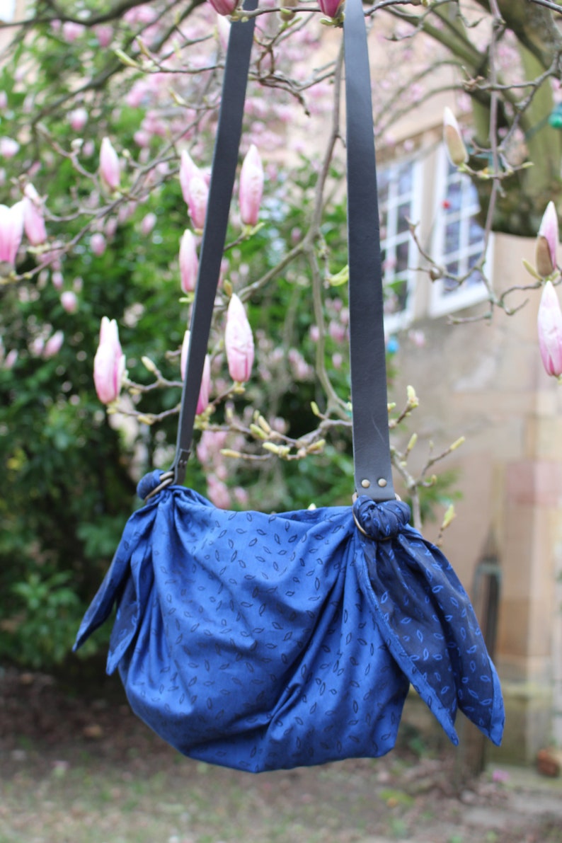 Puede incluir: Una bolsa de tela azul con un patr&oacute;n de hojas que cuelga de correas de cuero negro. La bolsa est&aacute; atada en la parte superior con un nudo y tiene un aspecto informal.
