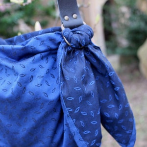 Puede incluir: Una envoltura de beb&eacute; de tela azul con un patr&oacute;n de hojas. La envoltura est&aacute; atada con un anillo y tiene correas de cuero.