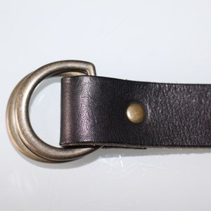 Puede incluir: Un cintur&oacute;n de cuero negro con una hebilla de doble anillo y un remache de lat&oacute;n. El cintur&oacute;n est&aacute; hecho de cuero de alta calidad y tiene un dise&ntilde;o cl&aacute;sico.