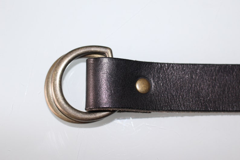Puede incluir: Un cintur&oacute;n de cuero negro con una hebilla de doble anillo y un remache de lat&oacute;n. El cintur&oacute;n est&aacute; hecho de cuero de alta calidad y tiene un dise&ntilde;o cl&aacute;sico.