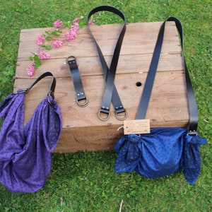 Puede incluir: Dos bolsas de tela de color morado y azul con correas de cuero negro y anillos de metal. Las bolsas est&aacute;n sobre una superficie de madera con algunas flores rosas.