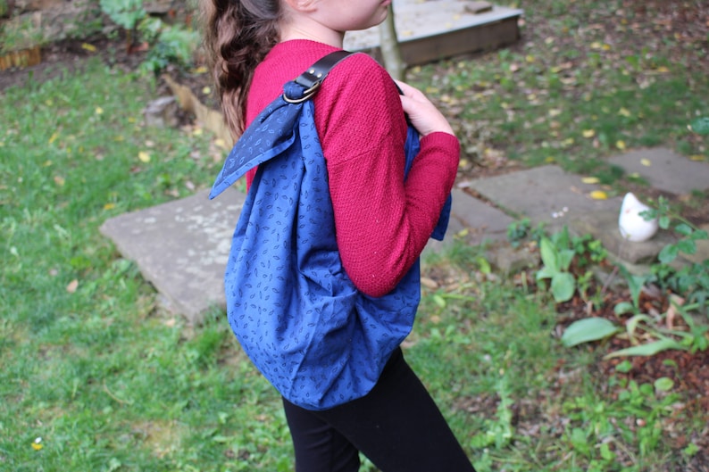 Puede incluir: Un bolso tote azul y negro con estampado y una correa de cuero marr&oacute;n. El bolso se lleva sobre el hombro de una persona que lleva un su&eacute;ter rojo y pantalones negros.