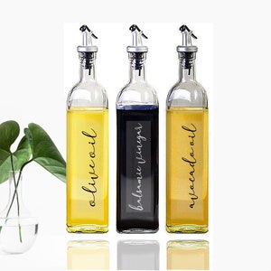 Puede incluir: Tres botellas de vidrio con boquillas de vertido y etiquetas. Las botellas est&aacute;n llenas de l&iacute;quidos. Las etiquetas dicen "aceite de oliva", "vinagre bals&aacute;mico" y "aceite de aguacate".