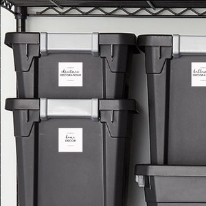 May include: Three black plastic storage bins with white labels. The top bin is labeled "Christmas Decorations", the middle bin is labeled "Home Decor", and the bottom bin is labeled "Halloween Decorations".