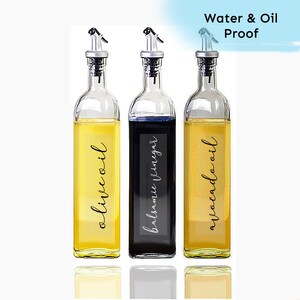 Puede incluir: Tres botellas de vidrio con etiquetas negras y boquillas negras. Las etiquetas dicen "aceite de oliva", "vinagre bals&aacute;mico" y "aceite de aguacate". Las botellas est&aacute;n sobre una superficie blanca. A prueba de agua y aceite.