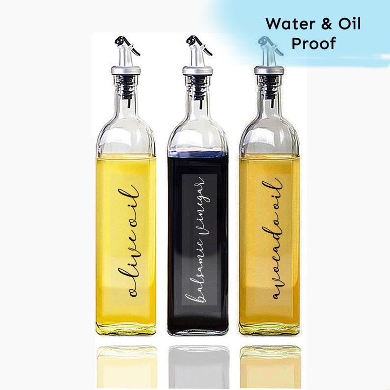 Puede incluir: Tres botellas de vidrio con etiquetas negras y boquillas negras. Las etiquetas dicen "aceite de oliva", "vinagre bals&aacute;mico" y "aceite de aguacate". Las botellas est&aacute;n sobre una superficie blanca. A prueba de agua y aceite.