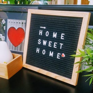 May include: A square felt letter board with a light wood frame and a black grooved background. White plastic letters spell out the phrase HOME SWEET HOME, accompanied by a small white arrow icon and a tiny red heart symbol on the board surface.