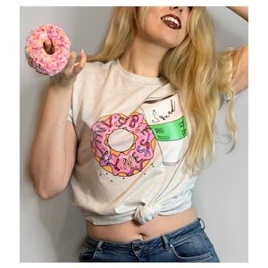 Peut inclure: T-shirt gris clair avec un motif de donut rose et l'inscription "Vibes & Sweetness", ainsi qu'une tasse de café avec "Squad". Un donut rose et jaune est tenu en l'air.