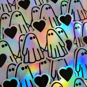 May include: A collection of iridescent holographic stickers featuring ghosts and hearts. The stickers are black and white with a rainbow iridescent finish.