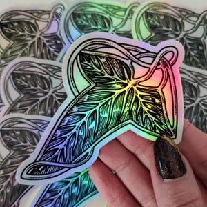 May include: A holographic sticker featuring a black and white illustration of a leaf with a swirling vine design. The sticker has a rainbow iridescent finish.