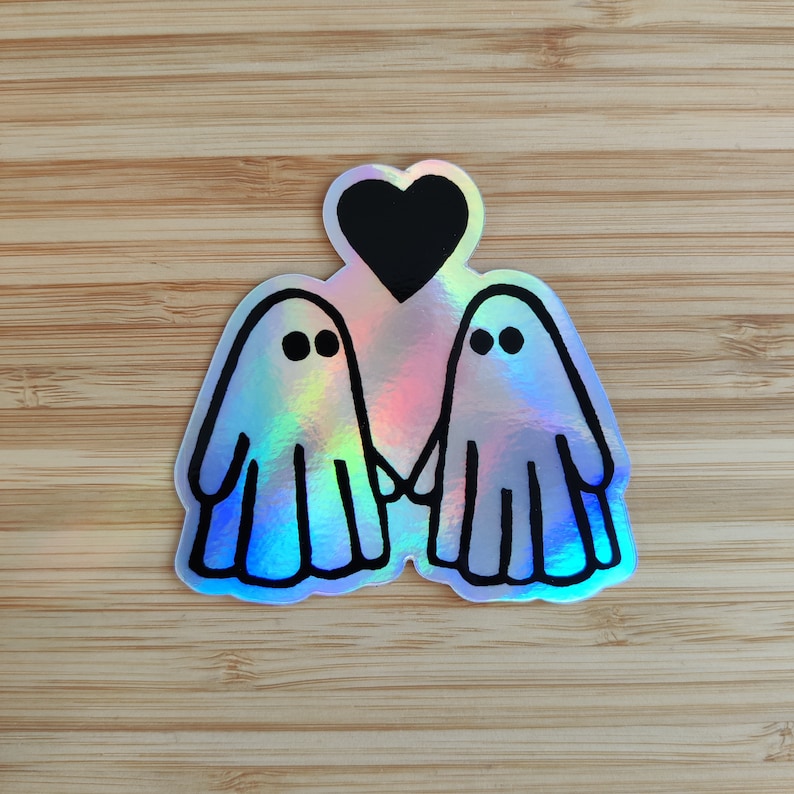 May include: A holographic sticker featuring two ghosts holding hands with a black heart above them. The ghosts are outlined in black and have simple eyes.