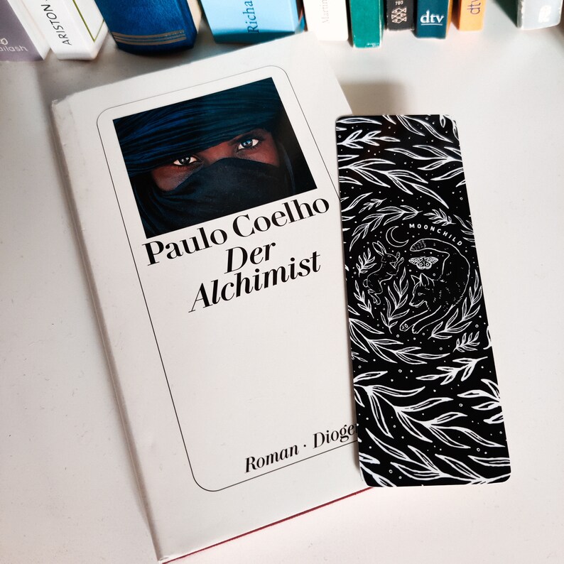 May include: A white book with the title "Paulo Coelho Der Alchemist" and the text "Roman Diogenes" on the bottom. The book is open and a black and white bookmark with a floral design and the text "Moonchild" is sticking out.