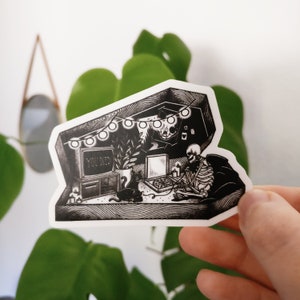 May include: Black and white illustration of a skeleton sitting in a coffin-shaped room, playing a video game with a cat sitting next to them. The game screen reads "YOU DIED". The room is decorated with string lights and a plant.