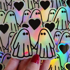 May include: A set of iridescent holographic stickers featuring two ghosts holding hands with a black heart between them. The stickers are surrounded by other ghost and heart stickers.