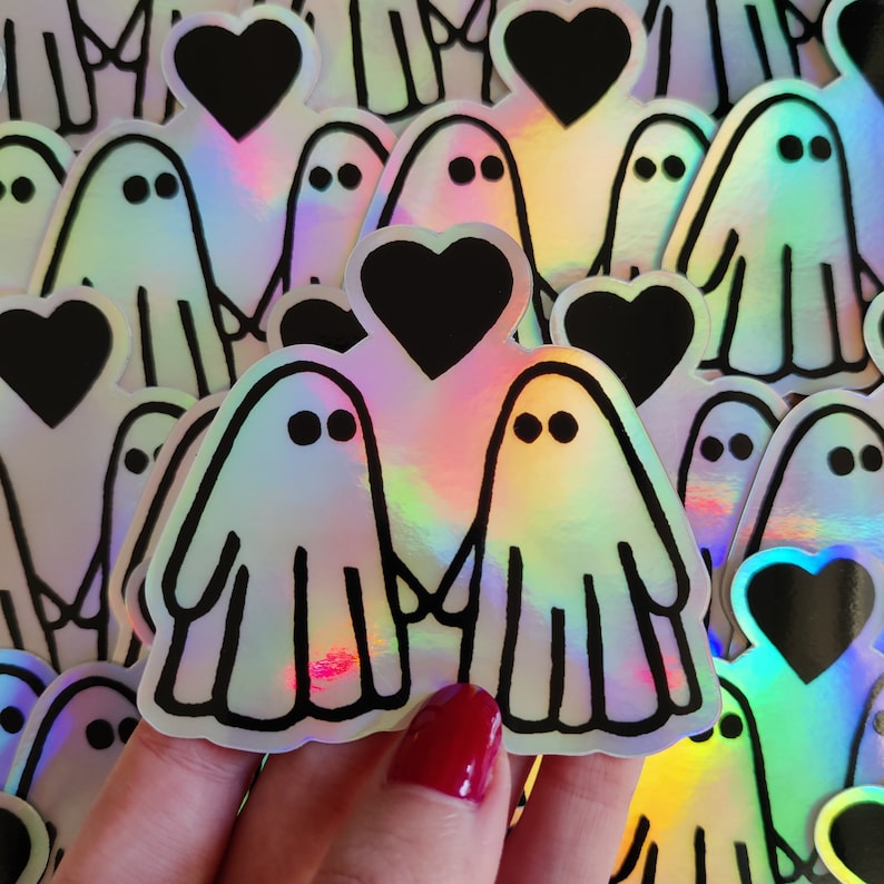 May include: A set of iridescent holographic stickers featuring two ghosts holding hands with a black heart between them. The stickers are surrounded by other ghost and heart stickers.