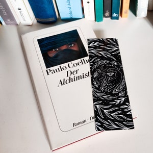 May include: A white book with a black and white bookmark. The book cover features a photo of a person wearing a blue headscarf and the text "Paulo Coelho Der Alchemist". The bookmark has a black background with white floral designs and the text "MOONCHILD".