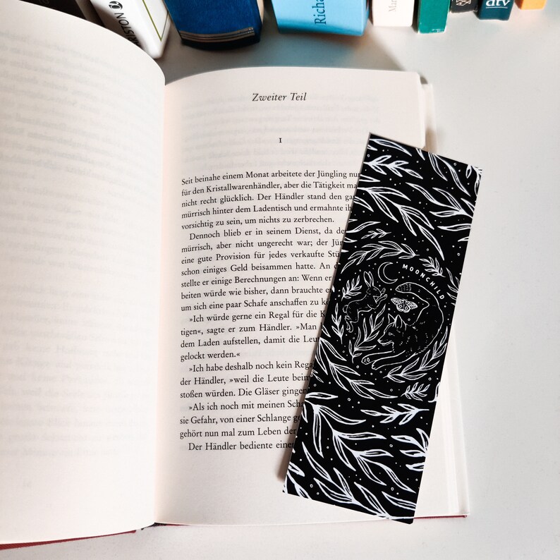 May include: A black and white bookmark with a whimsical illustration of a moon, a moth, and a creature with large ears and a long tail. The bookmark has the text "MOONCHILD" printed on it.