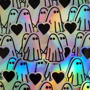 May include: A sheet of iridescent holographic stickers featuring ghosts holding hearts. The ghosts are outlined in black and have two eyes. The hearts are black.