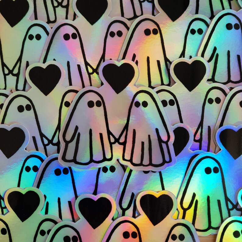 May include: A sheet of iridescent holographic stickers featuring ghosts holding hearts. The ghosts are outlined in black and have two eyes. The hearts are black.