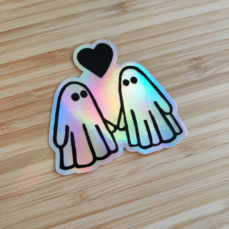 May include: A holographic sticker featuring two cartoon ghosts holding hands with a black heart above them. The ghosts are outlined in black and have black eyes.