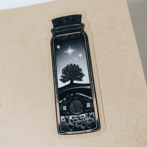 Könnte beinhalten: Eine Schwarzweißillustration einer Flasche mit einer Szene darin. Die Szene zeigt ein kleines Haus mit einer runden Tür, einen Baum und Sterne am Nachthimmel.
