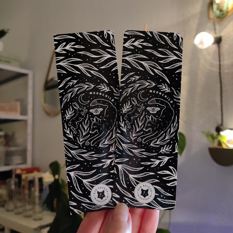 May include: Two black and white bookmark designs with a whimsical forest scene. The bookmarks feature a moon, a rabbit, a fox, a butterfly, and the text "Moonchild". The bottom of each bookmark has a small circle with the text "Kim Becker Design".