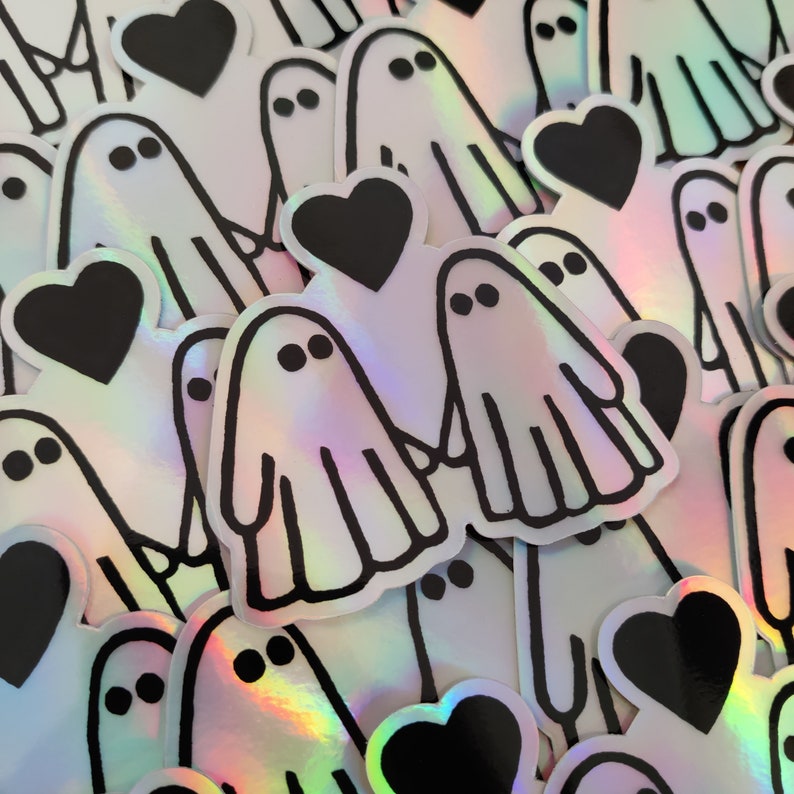 May include: A sheet of iridescent holographic stickers featuring a ghost holding a heart. The stickers are black and white with a rainbow sheen.