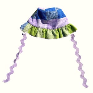 May include: A patterned bucket hat in shades of blue, purple, and green. The hat has a ruffled brim and wavy lavender ties. This handmade hat is a unique fashion accessory, perfect for adding a pop of colour.