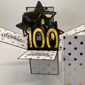 May include: A pop-up birthday card with a black and gold color scheme. The card features the number "100" in gold, surrounded by gold stars. The card is decorated with gold polka dots and the words "Celebrate" and "Happy Birthday".