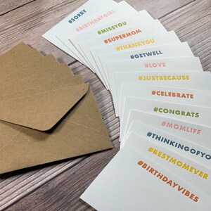 May include: A set of blank greeting cards with colourful hashtags printed on each card. The hashtags include #sorry, #birthdaygirl, #missyou, #supermom, #thankyou, #getwell, #love, #justbecause, #celebrate, #congrats, #momlife, #thinkingofyou, #bestmomever, and #birthdayvibes. The cards are stacked on top of each other and are surrounded by brown envelopes.