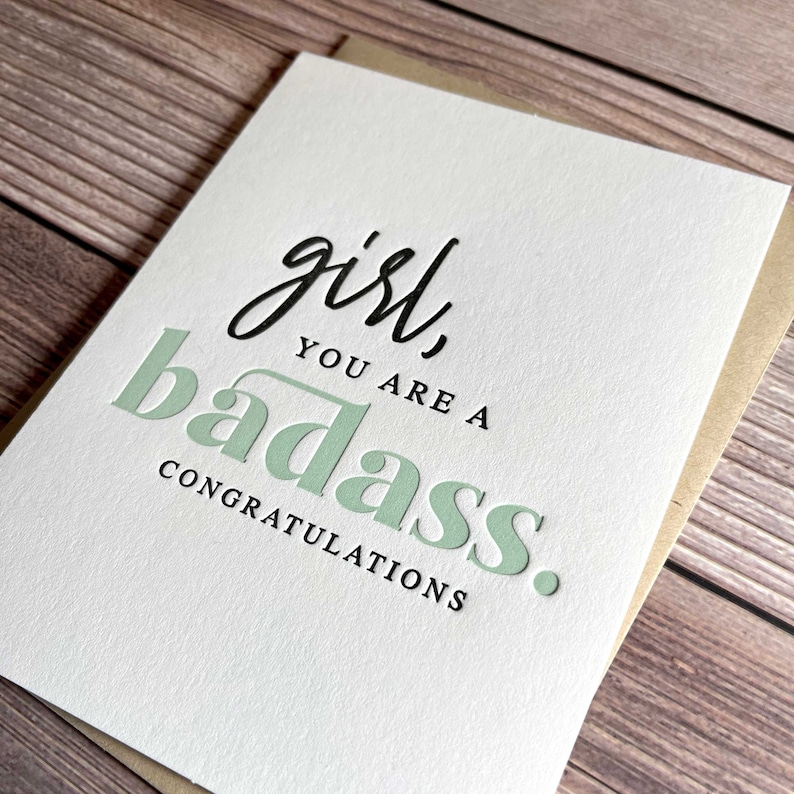 May include: A white card with the text "girl, you are a badass. Congratulations." in black and green lettering.