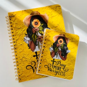 May include: Two yellow spiral bound journals with a black and white illustration of a woman holding a sunflower. The smaller journal has the text "Pray More Worry Less" on the front.