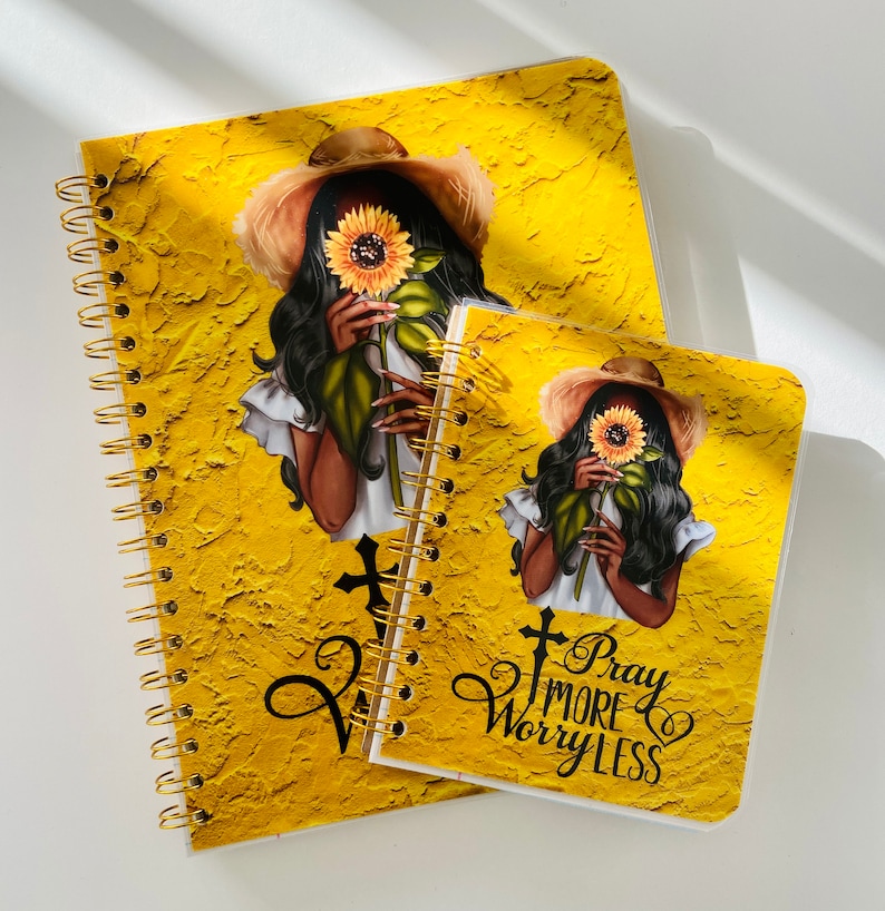 May include: Two yellow spiral bound journals with a black and white illustration of a woman holding a sunflower. The smaller journal has the text "Pray More Worry Less" on the front.