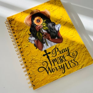 May include: A yellow spiral bound notebook with a textured surface. The cover features a black woman with a sunflower in her hand and the text "Pray More Worry Less".