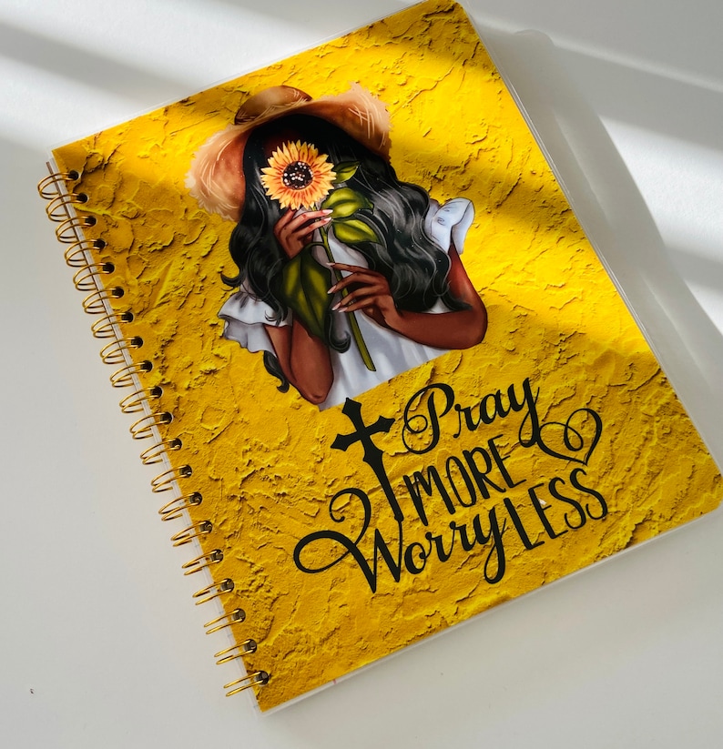May include: A yellow spiral bound notebook with a textured surface. The cover features a black woman with a sunflower in her hand and the text "Pray More Worry Less".