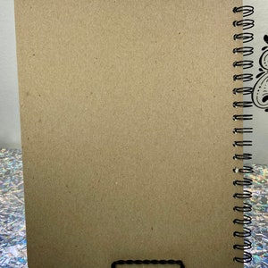 May include: A brown notebook with a black spiral binding. The notebook is sitting on a silver and iridescent surface.