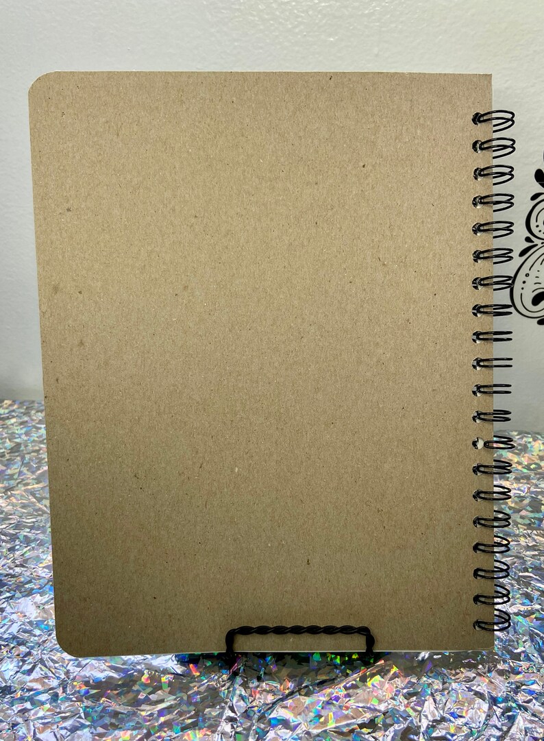 May include: A brown notebook with a black spiral binding. The notebook is sitting on a silver and iridescent surface.