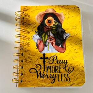May include: A yellow spiral bound notebook with a gold coil. The cover features a woman with long black hair wearing a straw hat and holding a sunflower. The text "Pray More Worry Less" is printed on the cover.