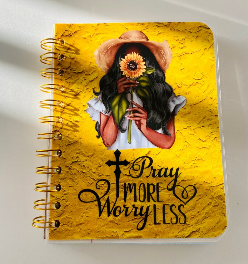 May include: A yellow spiral bound notebook with a gold coil. The cover features a woman with long black hair wearing a straw hat and holding a sunflower. The text "Pray More Worry Less" is printed on the cover.