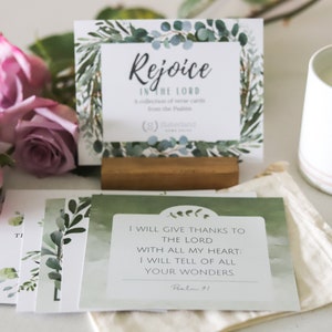 May include: A set of green and white cards with inspirational quotes from the Psalms. The cards are arranged in a stack with a wooden stand and a small white candle in the background. The text on the top card reads "Rejoice in the Lord".