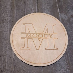 May include: A round wooden sign with the letter "M" and the name "MOODY" engraved in a light brown color. The sign is circular with a thin border and sits on a gray wooden surface.