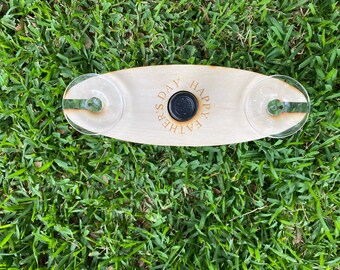 May include: A wooden wine glass holder with two slots for glasses and a center space for a bottle. The holder has the words "HAPPY FATHER'S DAY" engraved on it. The holder is sitting on green grass.