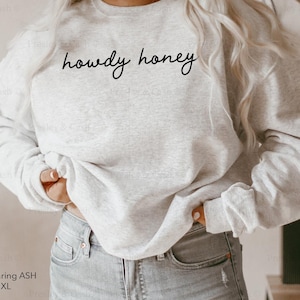 Pode incluir: Uma camisa de moletom cinza com o texto "howdy honey" em letras cursivas pretas.