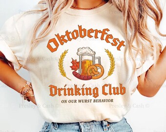 May include: Ivory-colored t-shirt with the text "Oktoberfest Drinking Club" in orange. The design includes a beer stein, pretzel, sausage, and wheat stalks. The phrase "ON OUR WURST BEHAVIOR" is below the club name.