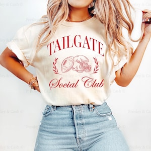 May include: A white t-shirt with the text "TAILGATE Social Club" in red lettering. The design features a football and helmet graphic. The shirt is being worn by a person with long blonde hair and blue jeans.